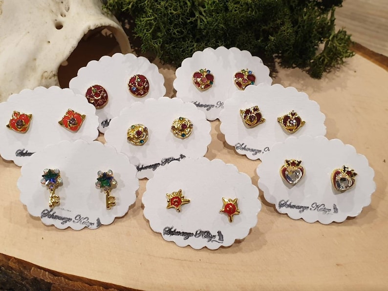May include: Assortment of colorful enamel earrings on white cards. The earrings feature various designs, including hearts, stars, keys, and crowns. Each card has the text "Adoraway. K" printed on it.