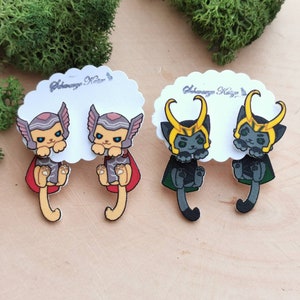 Pode incluir: Dois pares de brincos de gato, um par com um gato cinza usando um capacete vermelho e dourado e o outro par com um gato cinza usando um capacete verde e dourado.