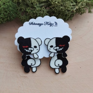 Peut inclure: Une paire de boucles d'oreilles en forme d'ours noir et blanc avec des yeux rouges. Les ours sont divisés en deux, une moitié noire et l'autre moitié blanche. Les boucles d'oreilles sont sur une carte blanche avec le texte "Ashnaga Kaji" écrit dessus.