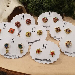 May include: A set of 10 pairs of earrings featuring various designs, including hearts, stars, and keys. Each pair is displayed on a white card with a letter label.