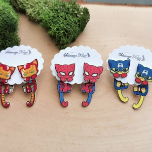 Może przedstawiać: Trzy pary kolczyków z superbohaterami w postaci kotów. Pierwsza para przedstawia Iron Mana, druga para przedstawia Spidermana, a trzecia para przedstawia Kapitana Amerykę.