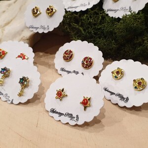 May include: A set of six pairs of gold-tone earrings with colorful enamel and rhinestones. The earrings are displayed on white cards with the text "Alchemy Key" printed on them.