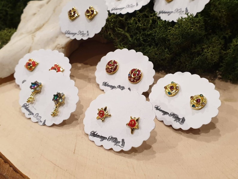 May include: A set of six pairs of gold-tone earrings with colorful enamel and rhinestones. The earrings are displayed on white cards with the text "Alchemy Key" printed on them.