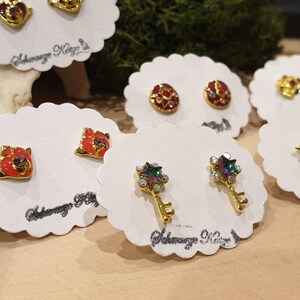 May include: Assortment of small, colorful, gold-tone earrings displayed on white cards. The earrings feature various designs, including stars, keys, and flowers.