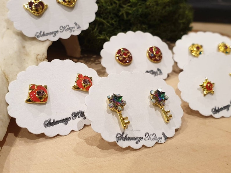 May include: Assortment of small, colorful, gold-tone earrings displayed on white cards. The earrings feature various designs, including stars, keys, and flowers.