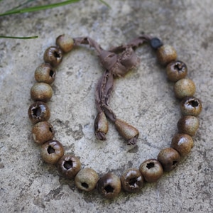 Puede incluir: Una pulsera con cuentas de cerámica redondas de color marrón moteado con hendiduras centrales, ensartadas en una cinta de tela marrón. Los extremos de la cinta terminan con dos pequeñas cuentas de cerámica a juego como adorno.