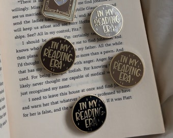 May include: Four enamel pins with the text "In My Reading Era" in different colors and fonts. The pins are on a book with text about a fictional character named Johanna.