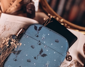 May include: A blue tablet case featuring a pattern of dragons, crowns, books, and stars. The case rests on an open book. A gold-framed mirror is visible in the background.