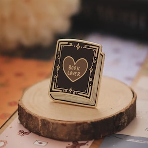 May include: A gold-coloured enamel pin in the shape of a book. The book has a black cover with a brown heart and the words "BOOK LOVER" inside. The pin is on a wooden slice.