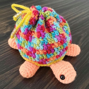 May include: A handmade crochet turtle-shaped pouch. The pouch is a vibrant mix of colors, including red, blue, green, and orange. It has peach-colored feet and a yellow drawstring. The turtle's eyes are black.