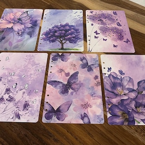 May include: Six planner inserts with floral and butterfly designs in shades of purple and pink. Each insert features a different watercolor illustration, including flowers, butterflies, and a tree. The inserts have pre-punched holes for binding.