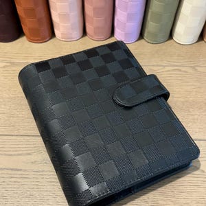 May include: A black personal planner with a textured checkerboard pattern and a matching snap closure strap. The planner is positioned in front of a row of similar planners in various colors including brown, pink, lavender, green, and cream.