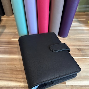 May include: A black personal planner with a textured finish and a snap closure strap, positioned in front of a row of upright planners in various colors including mint green, lavender, hot pink, beige, and deep purple with a cross-hatch texture.