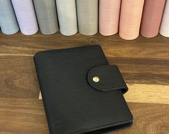 May include: A black leather-textured personal planner with a horizontal strap closure featuring a gold-tone metal snap button. The planner has a distinct horizontal ribbed pattern across its surface and is positioned in front of a row of similar planners in various pastel and neutral colors.