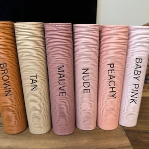 May include: A collection of six cylindrical items in various colors: brown, tan, mauve, nude, peachy, and baby pink. Each item has a textured surface and a label with its corresponding color name in black capital letters.