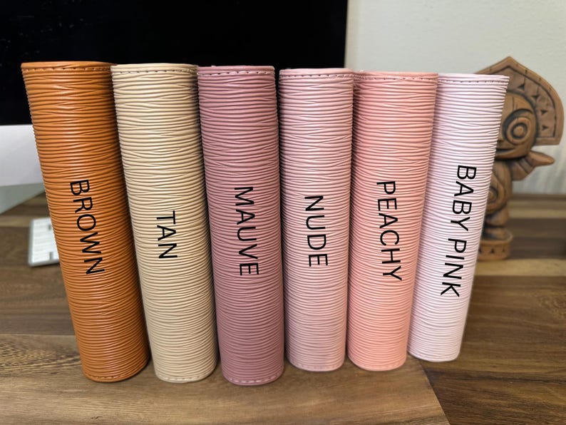 May include: A collection of six cylindrical items in various colors: brown, tan, mauve, nude, peachy, and baby pink. Each item has a textured surface and a label with its corresponding color name in black capital letters.