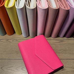May include: A collection of textured, envelope-style binders in a variety of colors including orange, yellow, white, beige, light pink, dusty rose, mauve, and lavender, with a vibrant hot pink binder placed prominently in the foreground.