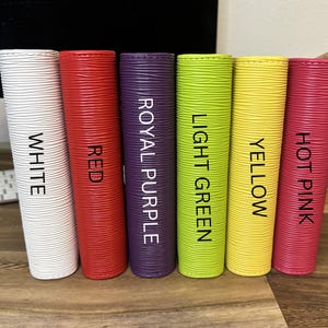 May include: Six cylindrical objects in white, red, royal purple, light green, yellow, and hot pink. Each has a textured surface and a label with its color name in black. The objects are arranged vertically on a wooden surface.
