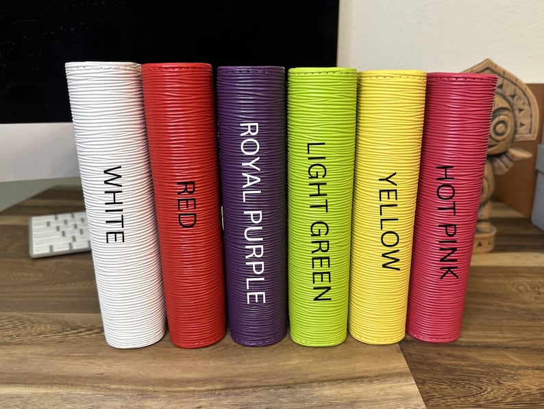 May include: Six cylindrical objects in white, red, royal purple, light green, yellow, and hot pink. Each has a textured surface and a label with its color name in black. The objects are arranged vertically on a wooden surface.