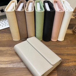 May include: A collection of eight rectangular planners or organizers with a textured lizard-skin pattern. Seven items stand upright in a row, while one light gray planner lies horizontally in the foreground. Each features a flap closure.