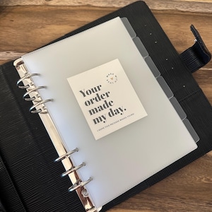 May include: Black leather binder with silver metal rings, holding a clear plastic sheet and a small card. The card reads "Your order made my day." and "I hope this package makes yours!"