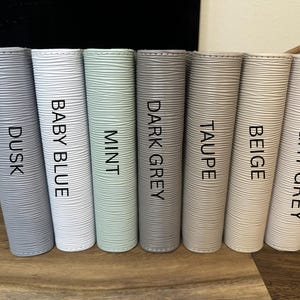 May include: Seven cylindrical containers in varying shades of gray, blue, and green are arranged in a row. Each container is labeled with a color name: Dusk, Baby Blue, Mint, Dark Grey, Taupe, Beige, and Light Grey. The containers have a textured surface.