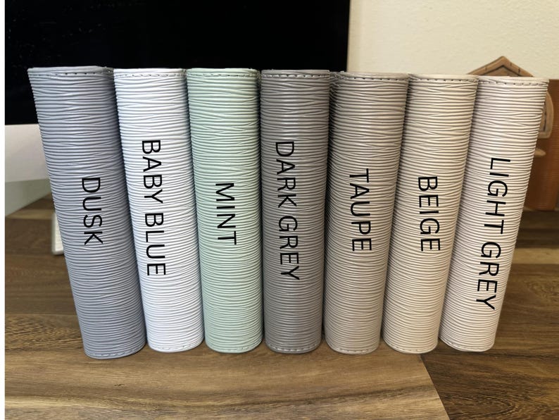 May include: Seven cylindrical containers in varying shades of gray, blue, and green are arranged in a row. Each container is labeled with a color name: Dusk, Baby Blue, Mint, Dark Grey, Taupe, Beige, and Light Grey. The containers have a textured surface.
