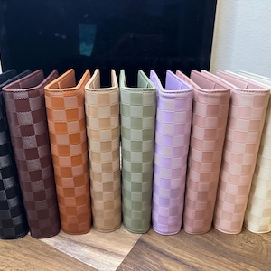 May include: A row of nine rectangular planners or notebooks standing upright, each featuring a textured checkerboard pattern. The colors include black, dark brown, tan, beige, sage green, lavender, dusty rose, light pink, and cream.