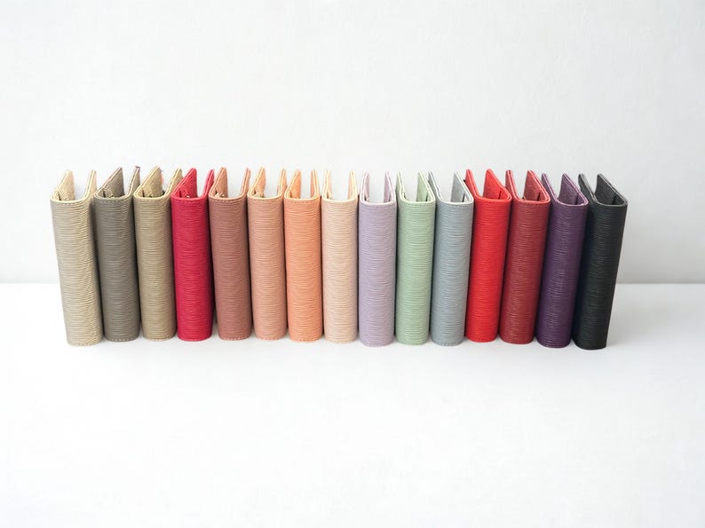 May include: A collection of colorful, textured passport holders in various shades, including beige, gray, red, and purple. The holders are arranged in a row against a white background, showcasing their diverse hues and designs.