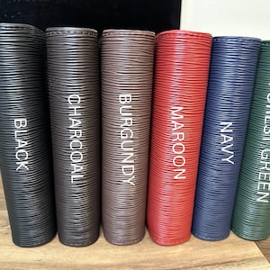 May include: Six vertical, textured leather-like items in various colors: black, charcoal, burgundy, maroon, navy, and forest green. Each item has a color name printed vertically in white. The items are arranged side by side on a wooden surface.