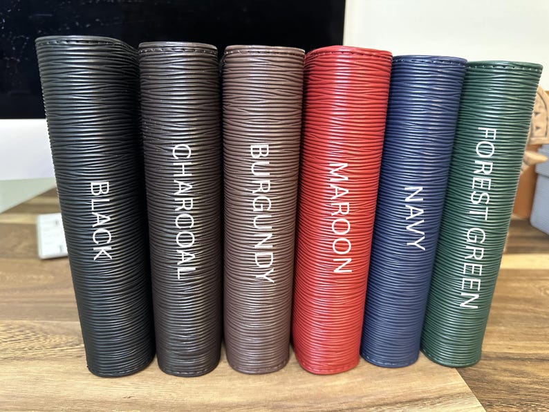 May include: Six vertical, textured leather-like items in various colors: black, charcoal, burgundy, maroon, navy, and forest green. Each item has a color name printed vertically in white. The items are arranged side by side on a wooden surface.