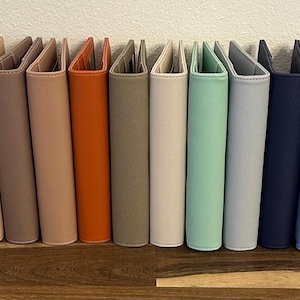 May include: A collection of colorful binders in various shades, including pink, burgundy, tan, orange, green, blue, purple, black, and silver. The binders are arranged in a row, with their spines facing the viewer, and are made of a smooth material.