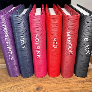 May include: A collection of six colorful, textured binders. The binders are in shades of royal purple, navy, hot pink, red, maroon, and black. Each binder has a white label with its color name. The binders are arranged on a wooden surface.