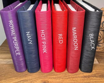 May include: A collection of six colorful, textured binders. The binders are in shades of royal purple, navy, hot pink, red, maroon, and black. Each binder has a white label with its color name. The binders are arranged on a wooden surface.