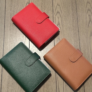 May include: Three rectangular planners with a textured, pebbled finish, each featuring a secure tab closure. The planners are presented in three distinct solid colors: a vibrant red, a deep forest green, and a warm, muted tan or light brown shade.