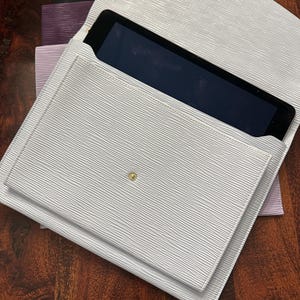 May include: A light gray, textured tablet case with a matching flap and gold snap closure. The case is open, revealing a black tablet screen. Additional cases in black, purple, and lavender are visible.