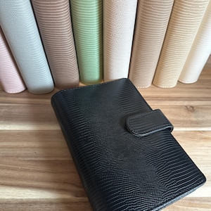 May include: A black, textured, faux-leather planner with a snap closure sits in front of a row of colorful planners. The planners are in shades of pink, gray, green, and beige. The background is a wooden surface.