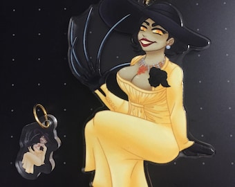 Pode incluir: Um grande chaveiro de acrílico amarelo com uma mulher sentada em um vestido amarelo e chapéu preto, segurando um guarda-sol preto. Um chaveiro menor com o mesmo design também está presente. Ambos têm ferragens douradas.