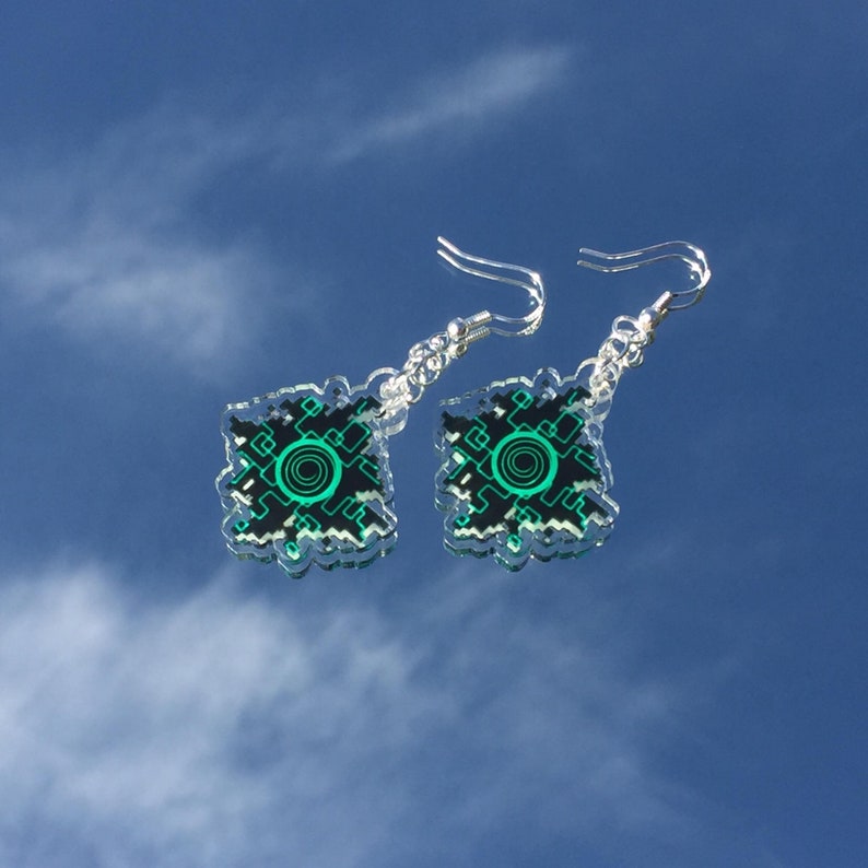 Pode incluir: Um par de brincos de prata com um design pixelado verde e preto. O design apresenta um c&iacute;rculo com um c&iacute;rculo menor dentro.