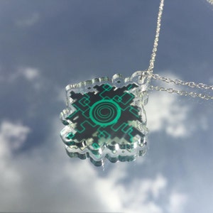 Pode incluir: Um colar de corrente prateada com um pingente de acr&iacute;lico transparente com um design geom&eacute;trico verde e preto. O design se assemelha a uma placa de circuito com um c&iacute;rculo verde no centro.