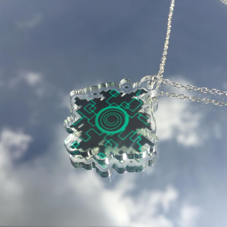 Pode incluir: Um colar de corrente prateada com um pingente de acr&iacute;lico transparente com um design geom&eacute;trico verde e preto. O design se assemelha a uma placa de circuito com um c&iacute;rculo verde no centro.