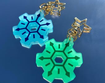 Pode incluir: Dois pingentes em forma de floco de neve azul e verde com detalhes de estrela dourada. Os pingentes estão presos a correntes douradas.