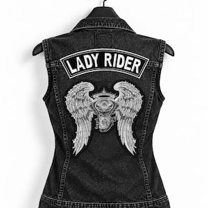 以下が含まれることがあります： 黒いデニムベスト。背中には「LADY RIDER」とアーチ状に刺繍された白いパッチがあり、その下にはバイクのエンジンと左右に大きく広がった羽が描かれた、詳細な刺繍パッチが施されています。