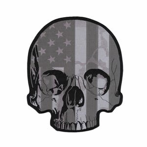 May include: A skull-shaped embroidered patch featuring a greyscale American flag pattern. The design incorporates black outlines for the eye sockets, nasal cavity, and teeth, with a thick black border surrounding the entire skull shape.