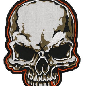 Pode incluir: Patch bordado de caveira em preto, branco e castanho, com contorno laranja e preto. O design da caveira apresenta sombreamento detalhado e um aspeto texturizado, ideal para personalizar roupas ou acessórios.