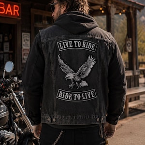 May include: A black denim jacket with a hood, featuring patches with the text "LIVE TO RIDE" and "RIDE TO LIVE" above and below an eagle graphic. The jacket is worn over a black hoodie and paired with black pants. A motorcycle is visible in the background.