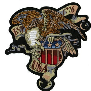 May include: Embroidered patch featuring a detailed eagle with outstretched wings, clutching a shield with stars and stripes. The patch includes the text "EST USA 1776" on banners, with a black border. The eagle has brown, tan, and white feathers.