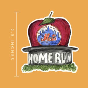 Puede incluir: Una manzana roja con una gorra de béisbol de los New York Mets en la parte superior. La manzana está sentada sobre una base negra con las palabras "Home Run" escritas en ella. La manzana tiene 2,5 pulgadas de alto.