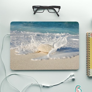 May include: A laptop computer with a beach scene on the cover. The scene shows a white rock with white foamy waves crashing around it on a sandy beach.