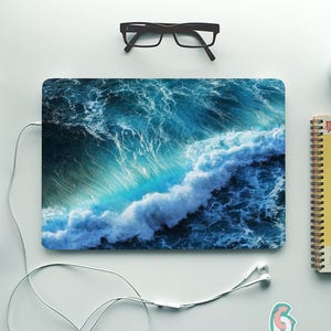 May include: A laptop computer with a blue and white ocean wave design on the cover. The laptop is on a white surface with a pair of glasses, a notebook, a pencil, and a cup of coffee.